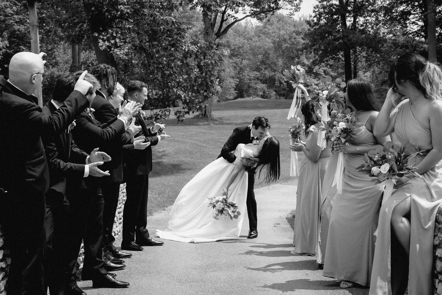 A groom dipping and kissing the bride on a bridge while the entire wedding party looks on and cheers.