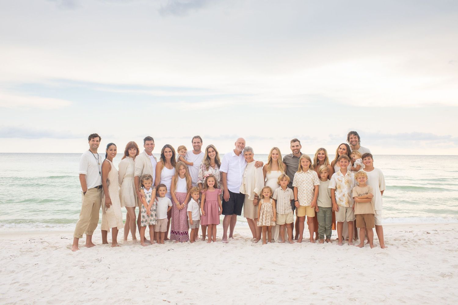 A group of people wearing coordinated beige and pastel outfits pose together on a sunny beach at sunset.