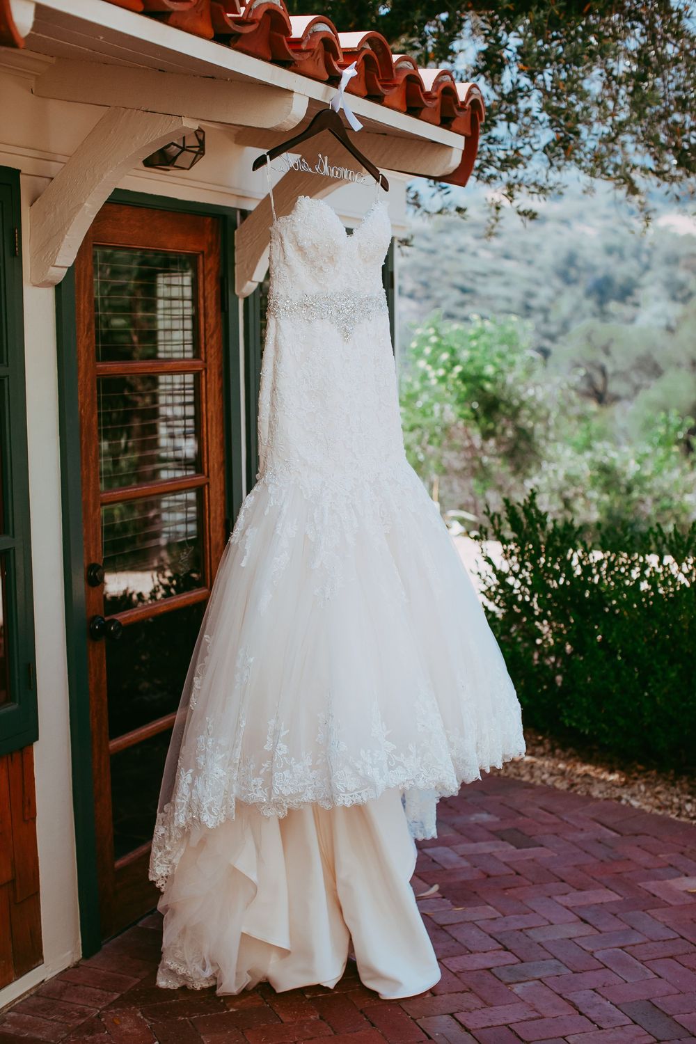 White wedding dress with delicate lace details hanging on display against rustic brick patio and garden backdrop.