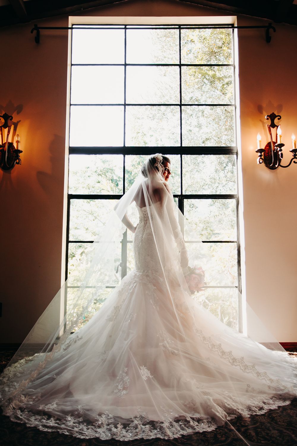 Elegant wedding dress with lace train silhouetted against large window with wall sconces creating dramatic lighting.