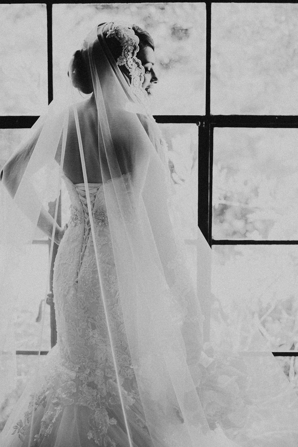 A bride in a flowing white wedding gown and long veil stands silhouetted against a bright window in black and white.