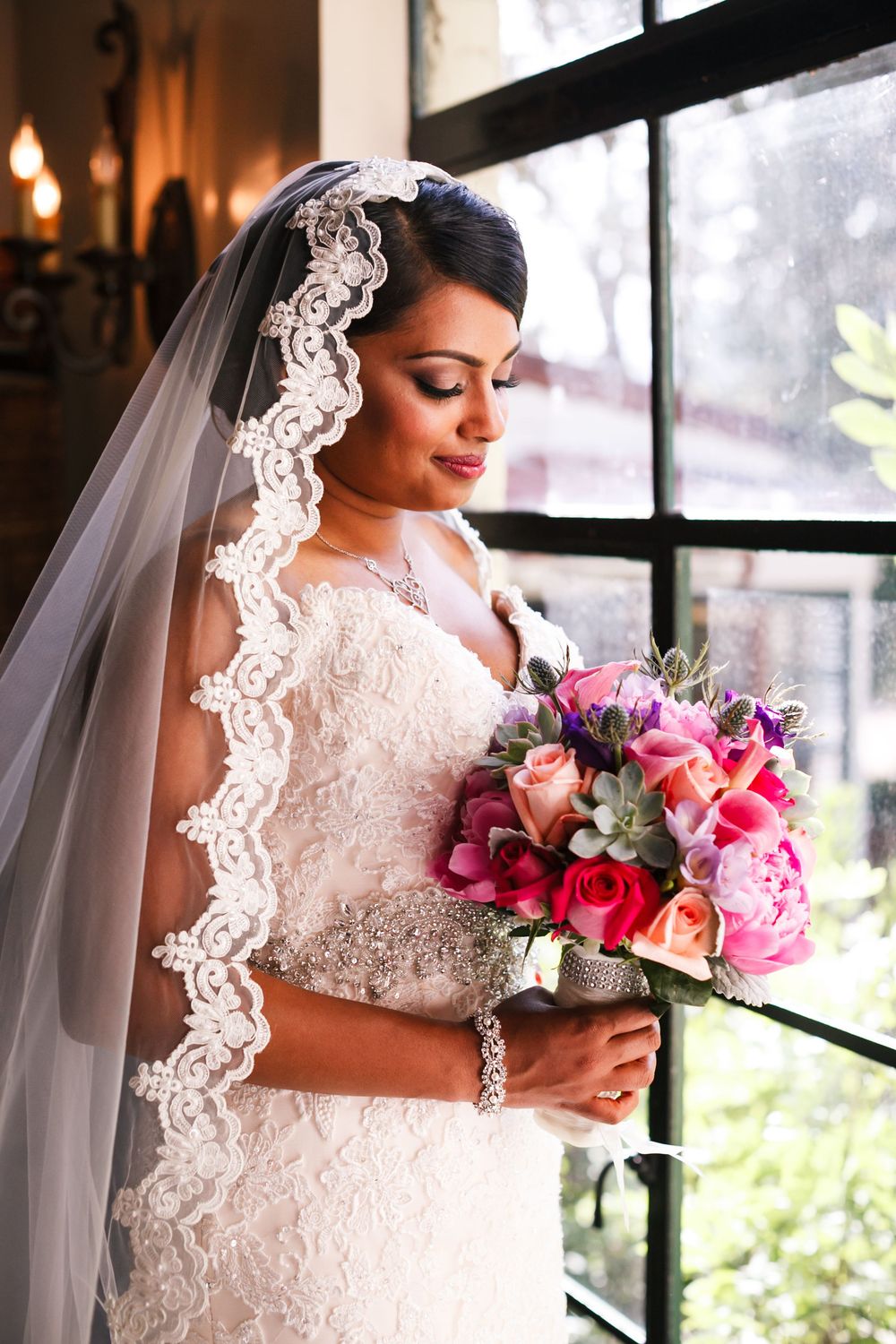 Bride in delicate lace wedding gown holds vibrant pink and purple bouquet while standing by sunlit window.