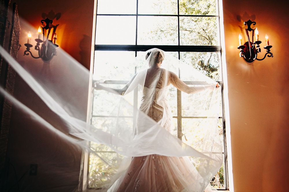 A wedding dress hangs in front of a sunlit window creating a romantic silhouette with warm lighting.