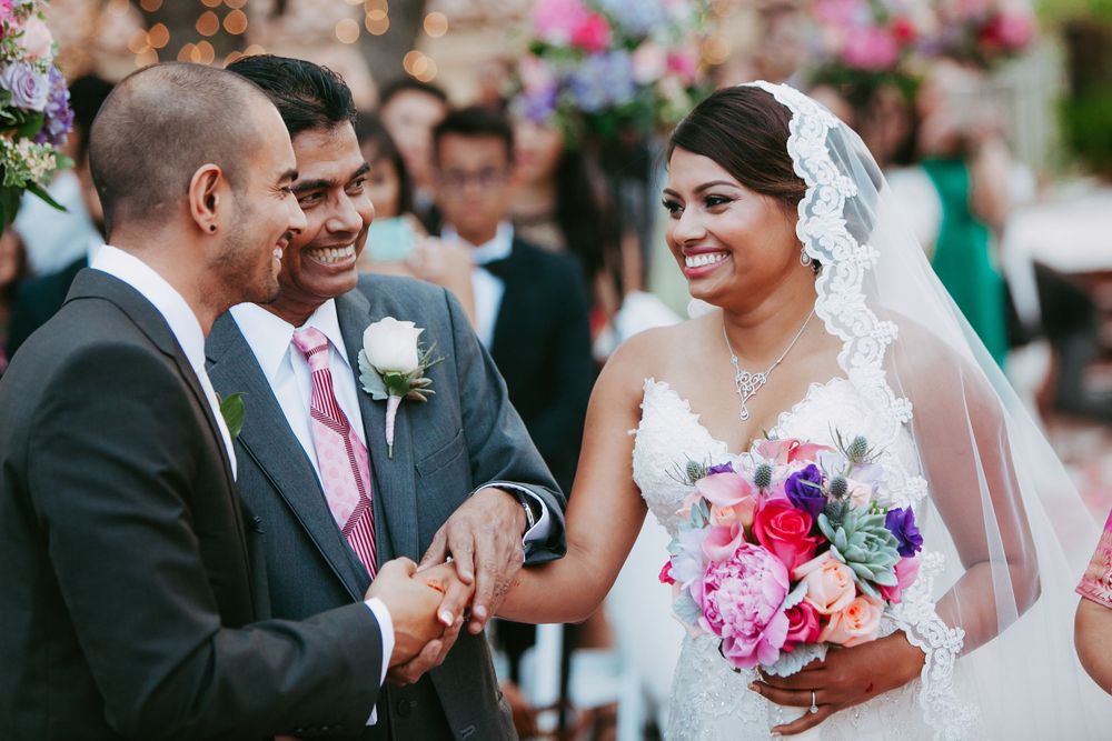Joyful wedding ceremony moment with couple exchanging rings as guests look on, featuring vibrant pink and purple bouquet.