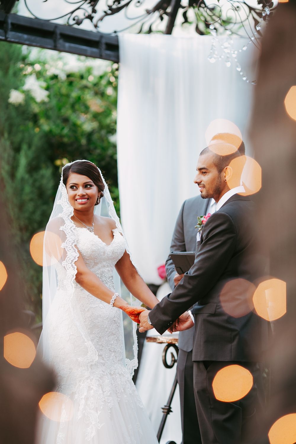 A wedding ceremony moment captured with soft bokeh lights and white drapery in an outdoor garden setting.
