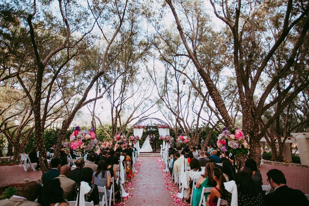Wedding guests toss pink flower petals during an outdoor ceremony beneath tall trees and a rustic wooden arch.