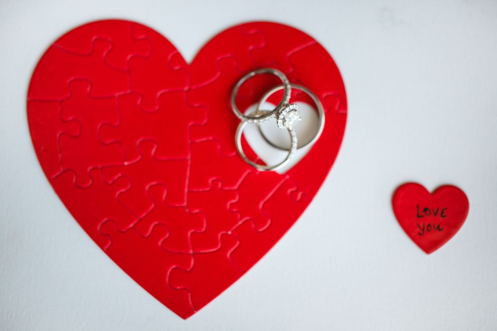 A silver wedding ring rests on a red heart-shaped jigsaw puzzle with a smaller heart in the background.