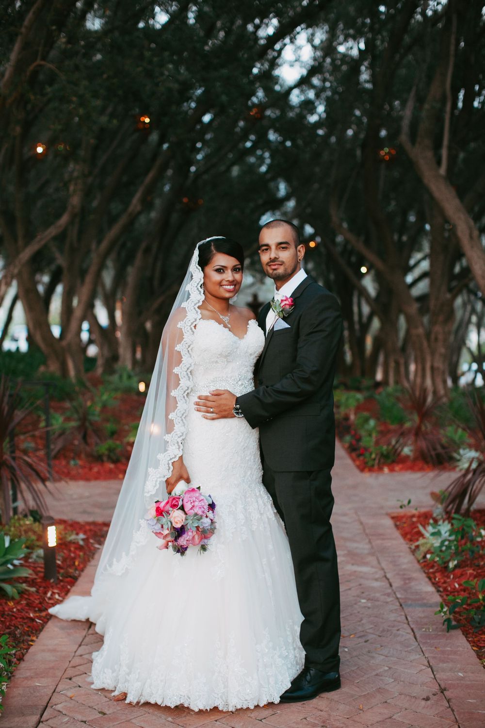 Bride and groom share romantic moments along a tree-lined path with red flowers during their wedding photoshoot.