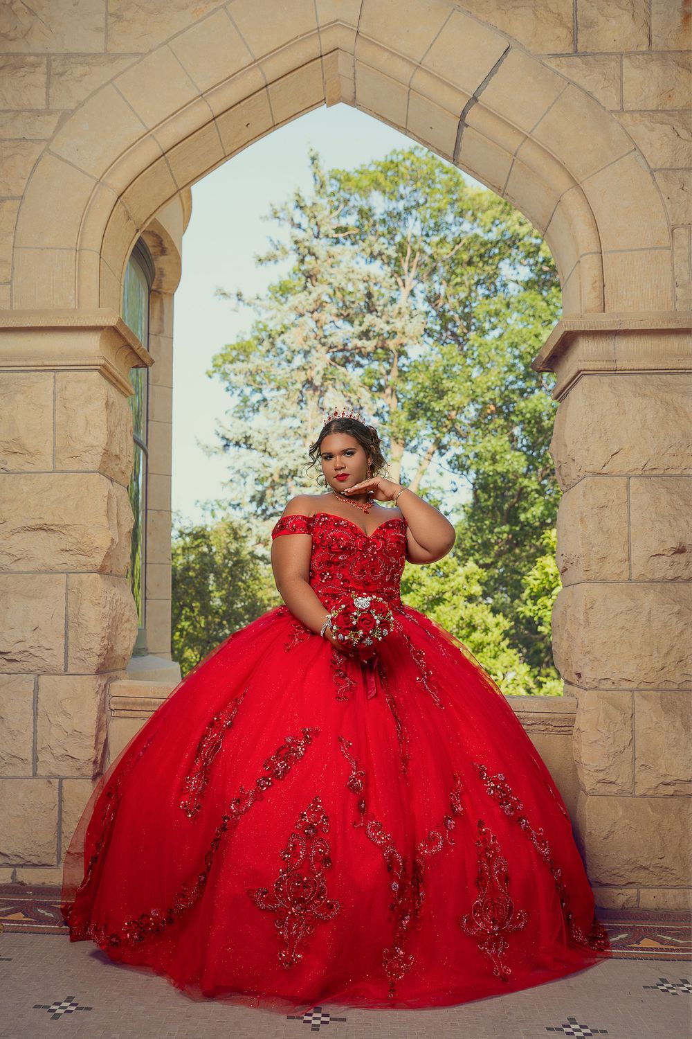 A stunning red quinceañera ball gown with intricate beading photographed against stone archways.