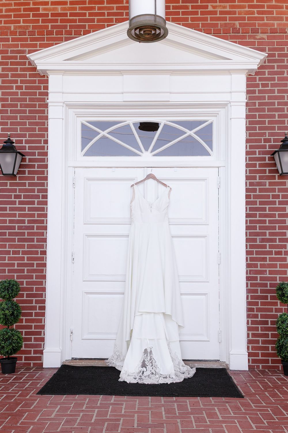 A white wedding dress hangs elegantly against a classic red brick wall with white door entrance.