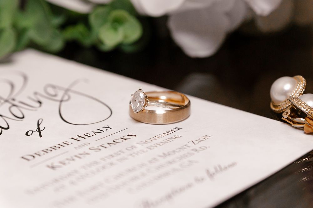 Wedding rings rest on a wedding invitation with elegant calligraphy text visible in soft lighting.