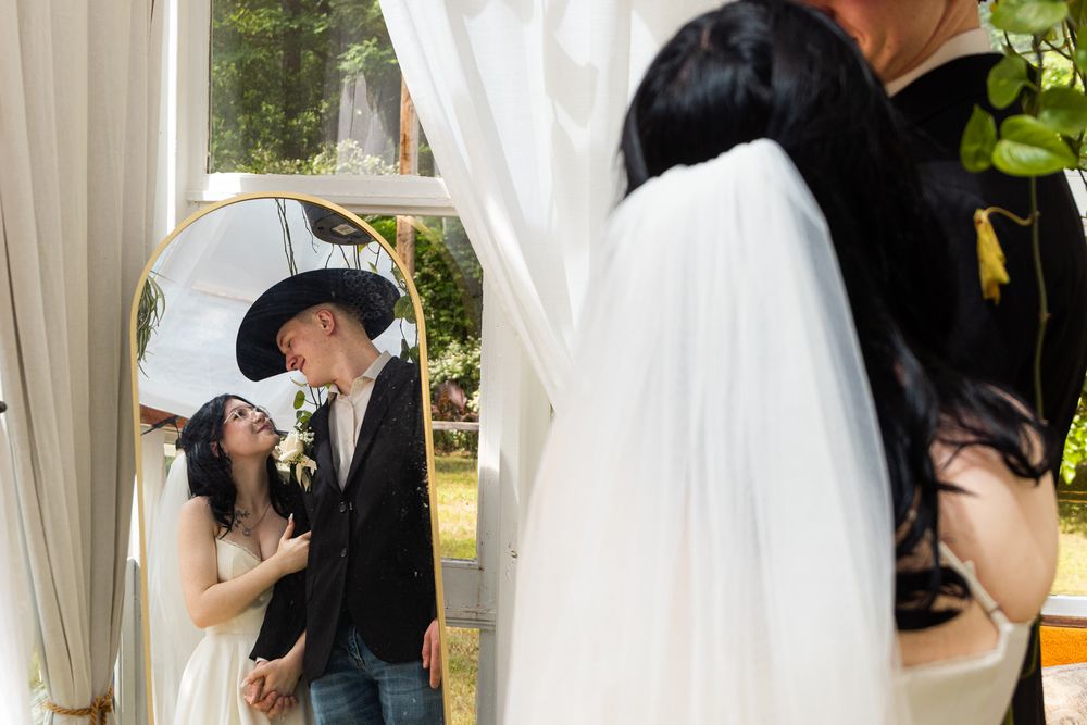 A romantic wedding couple shares a kiss under a white curtain arch while their reflection appears in a mirror beside them.