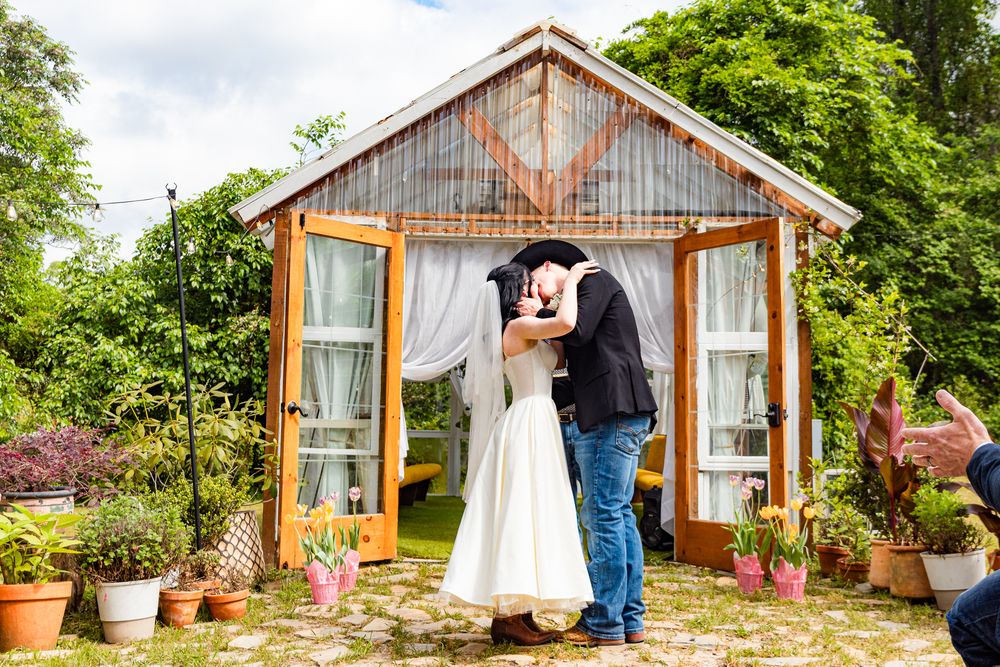 A rustic wooden greenhouse surrounded by lush greenery serves as a backdrop for a romantic outdoor moment.
