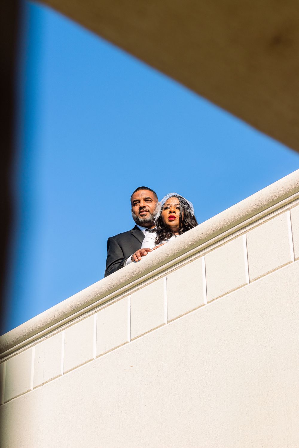 A couple shares a joyful moment together while looking down from a white balcony against a bright blue sky.
