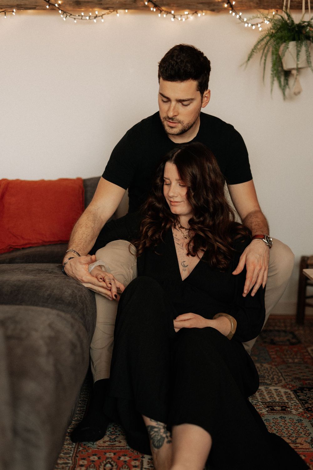 Couple in black and khaki clothing embraces on couch with string lights and decor in background.