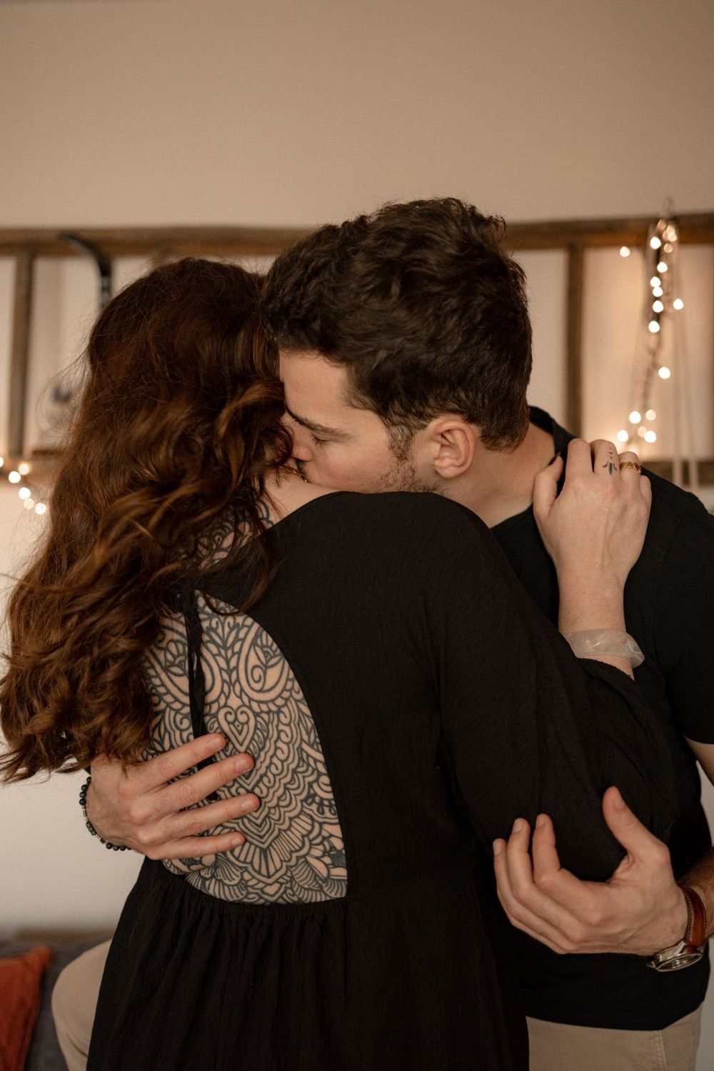A couple in dark clothing embraces intimately in a dimly lit room with string lights in the background.