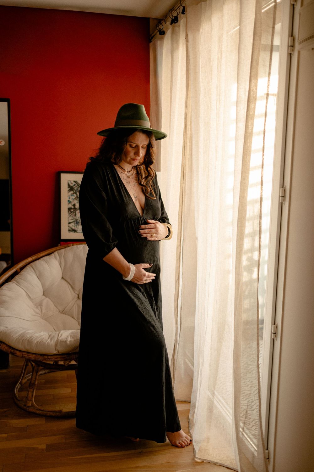 A person wearing a black dress and wide-brimmed hat stands by sheer curtains in a room with a red accent wall.