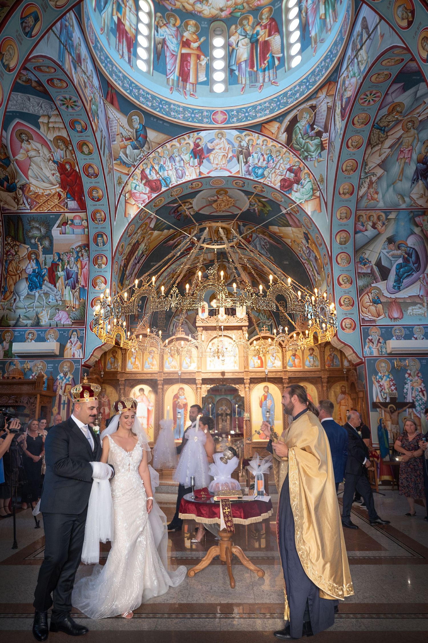 Bride and groom exchange vows in an ornate Orthodox church adorned with colorful frescoes and a grand chandelier.