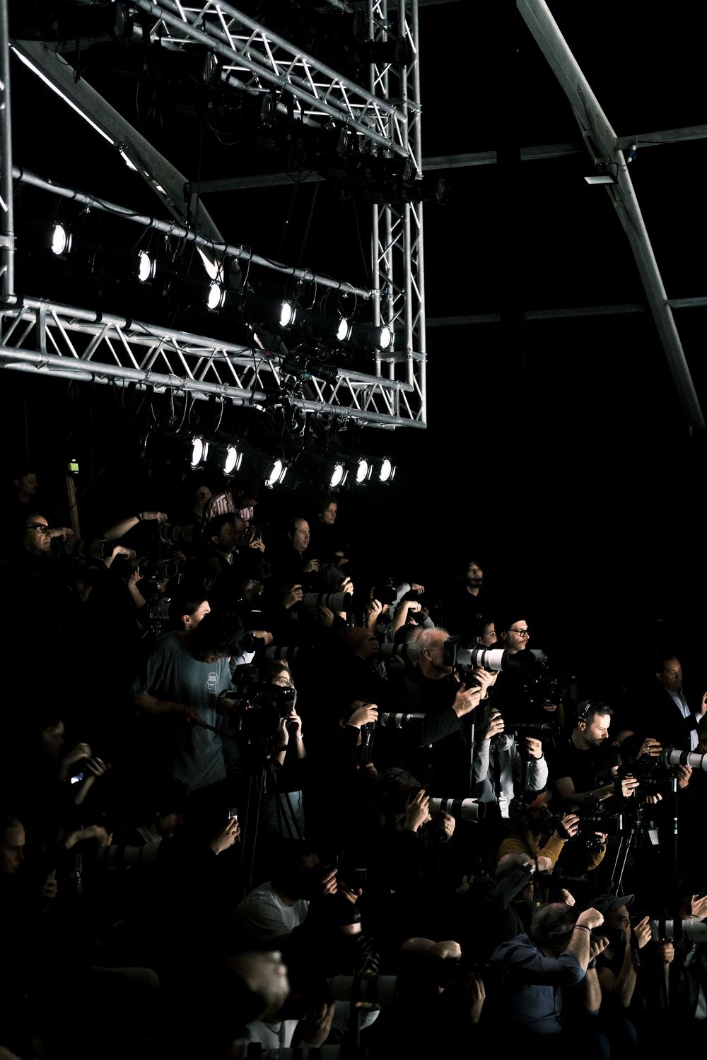 Photographers with cameras gather in a dark venue at a press event, capturing shots from multiple angles.