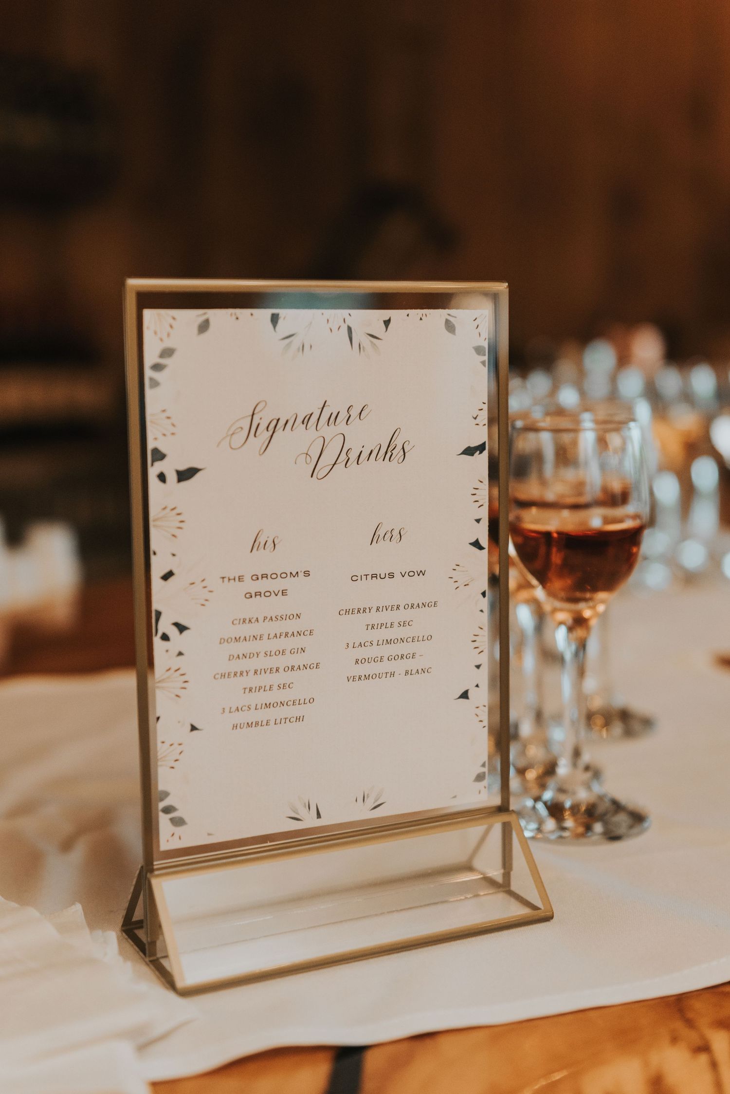 Elegant wedding signature drink menu displayed in a gold frame on white tablecloth with glassware in warm lighting.