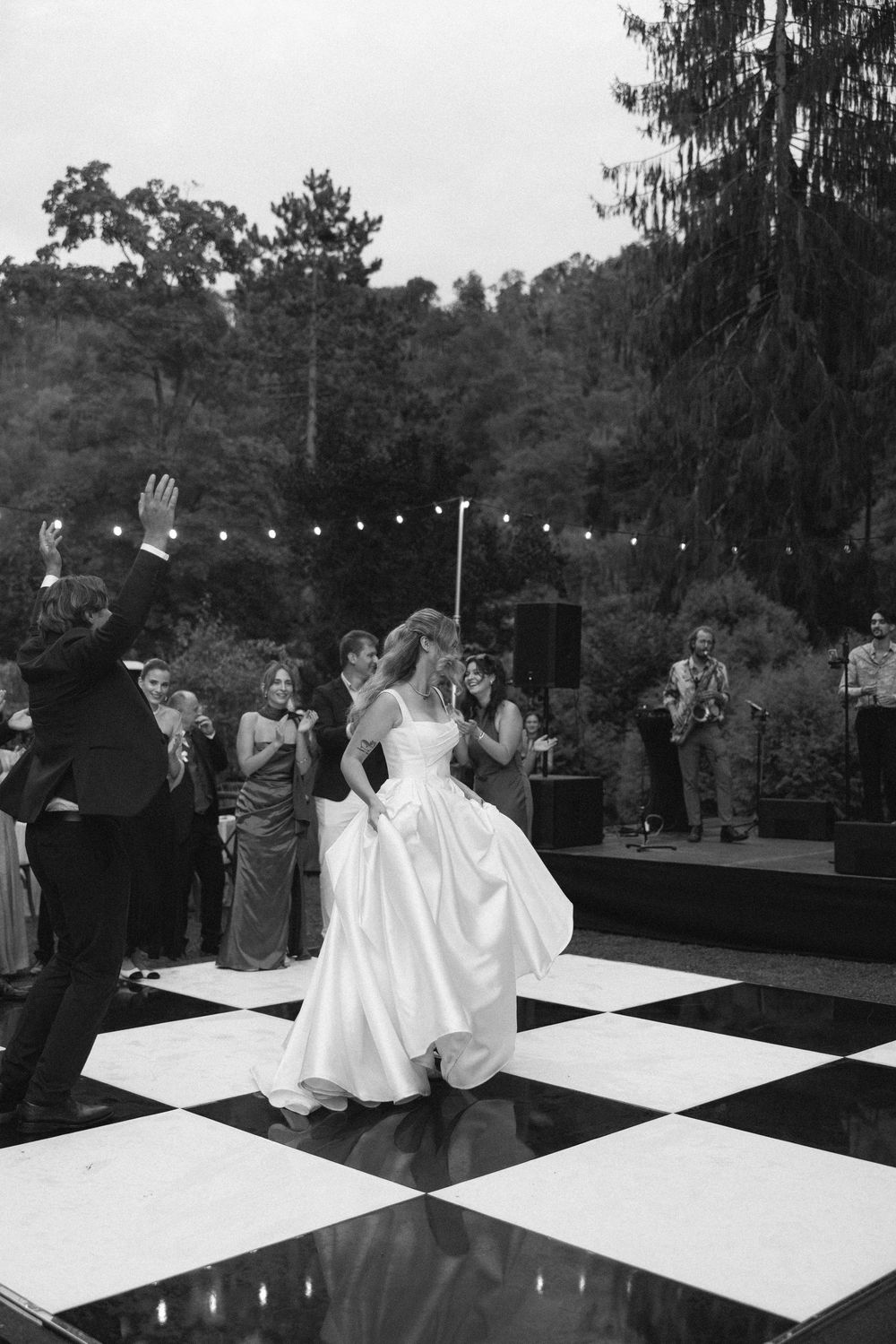 Wedding guests dance on a black and white checkered dance floor at an outdoor reception with string lights overhead.