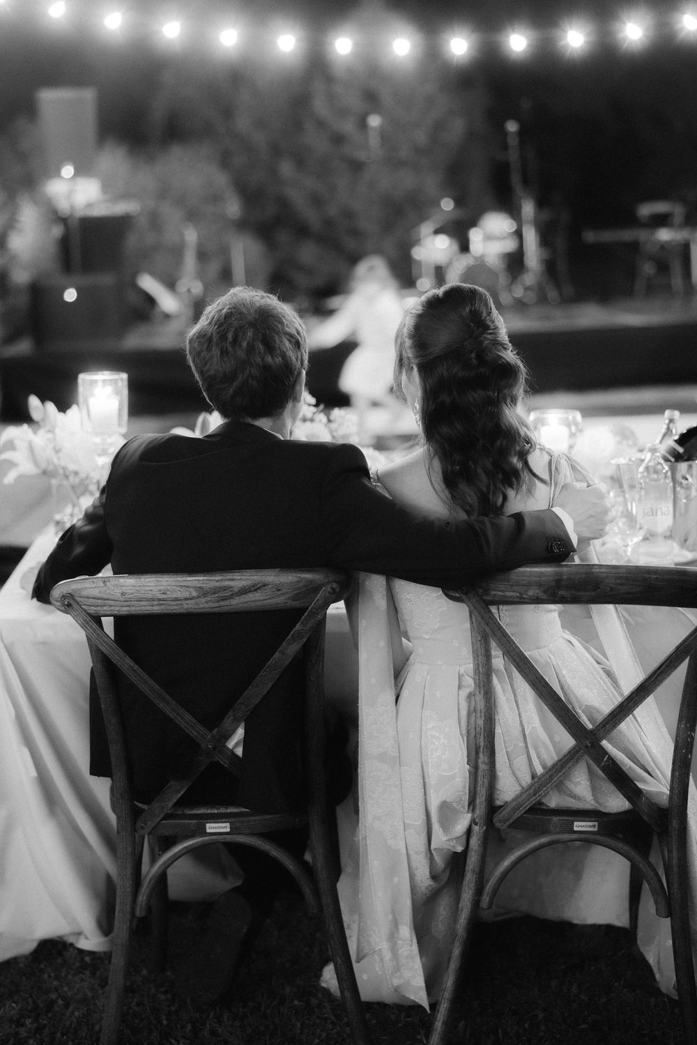 A couple sits together at an outdoor wedding reception under string lights in black and white photography.