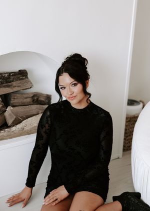 A person in a black long-sleeve sweater poses against a white wall with stacked firewood.