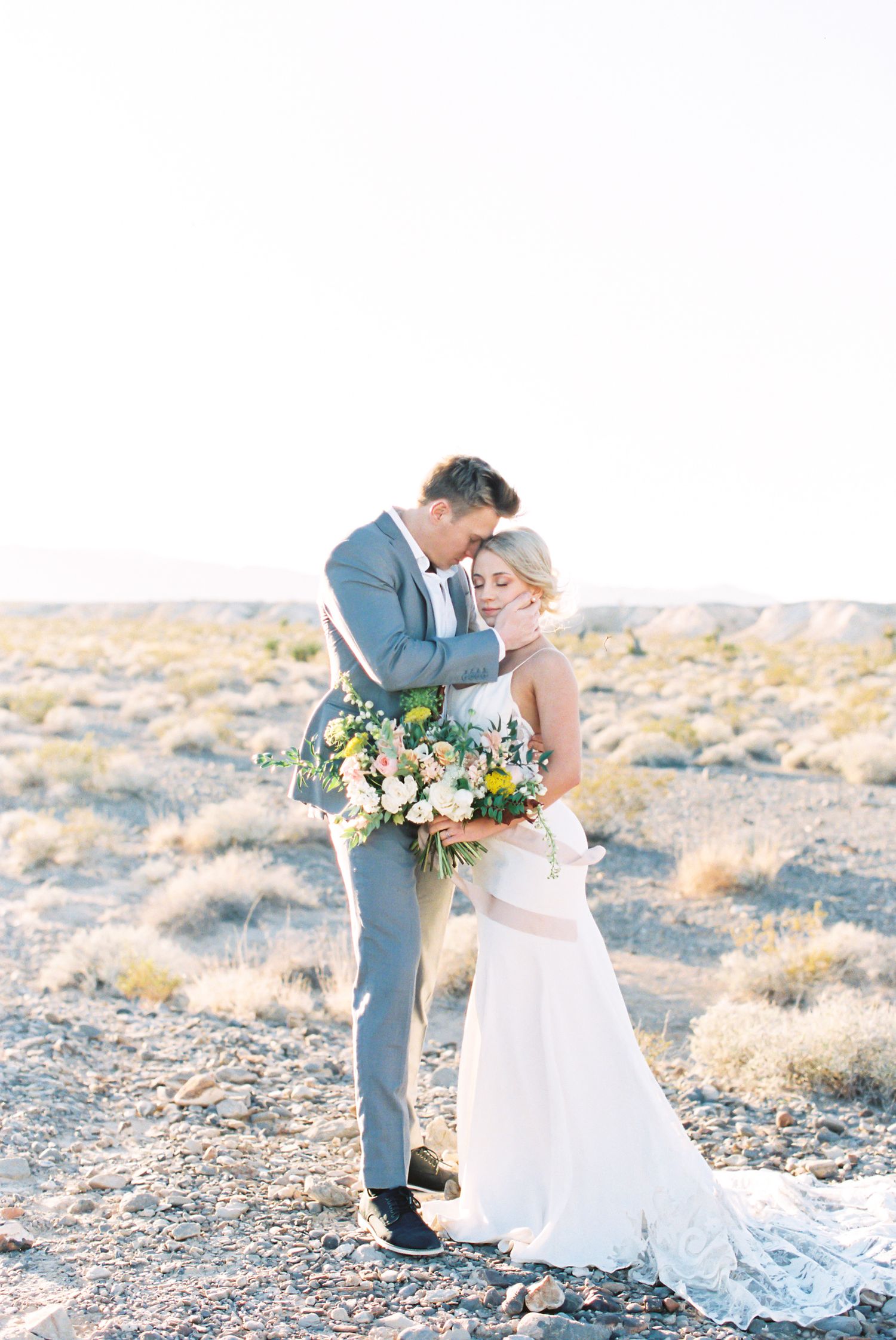 A romantic desert wedding photoshoot featuring a couple in formal attire embracing against a golden desert landscape.