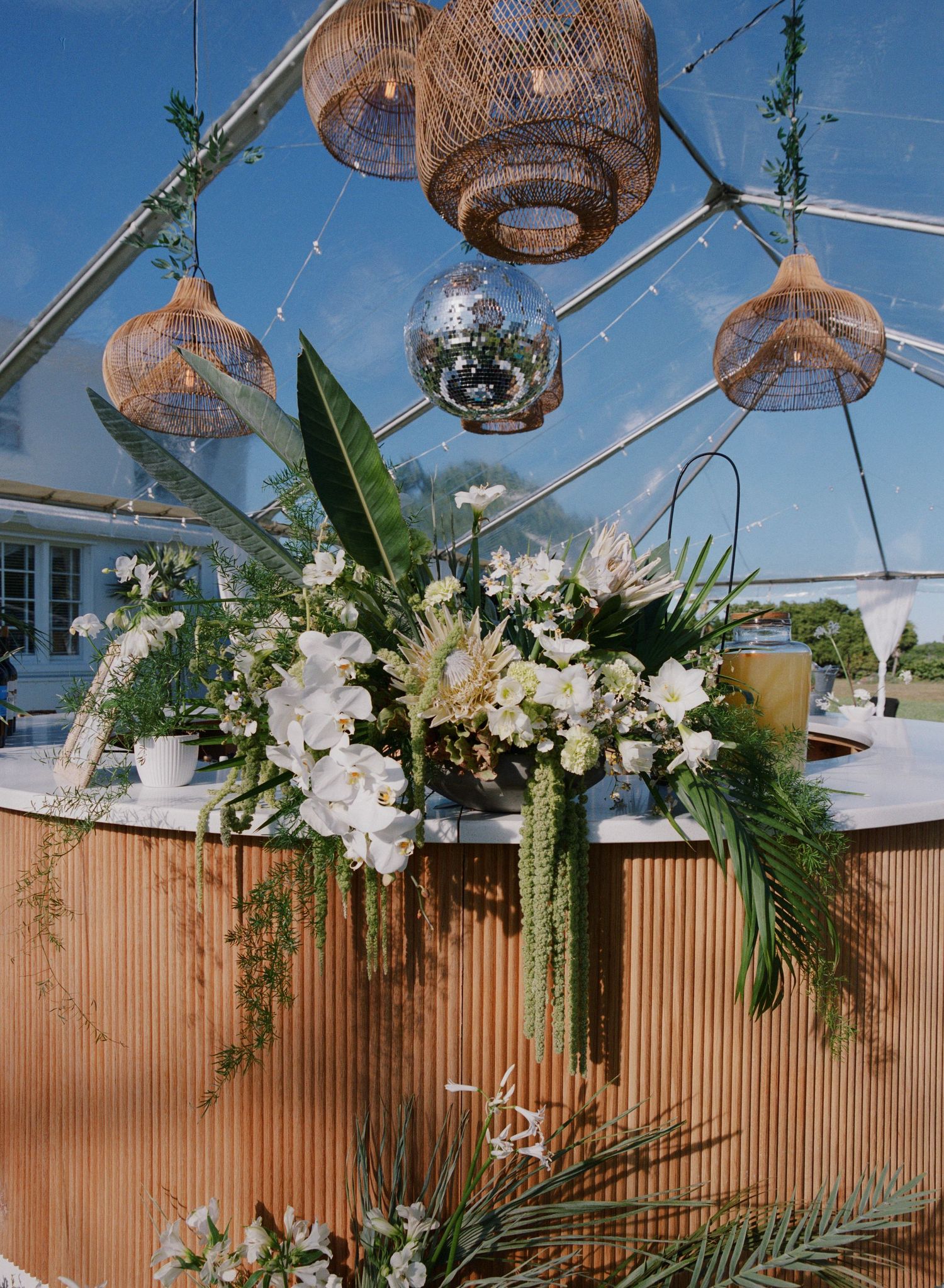 Elegant wedding reception decor with woven pendant lights, disco ball, and lush white floral arrangements under clear tent ceiling.