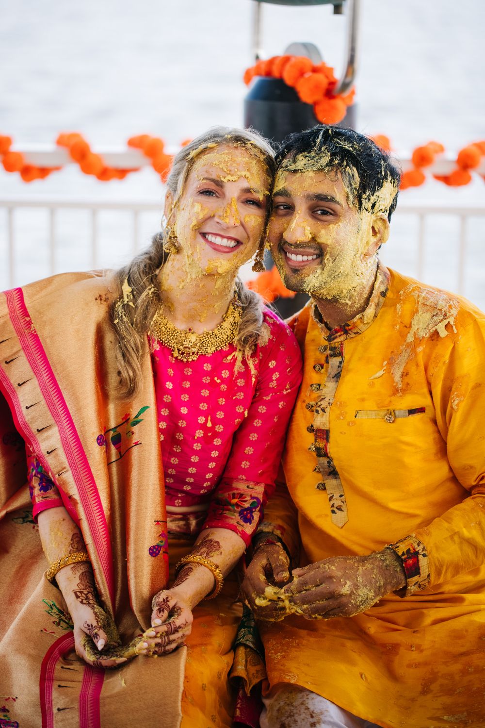 Joyful couple celebrates during a traditional Haldi ceremony with turmeric paste and bright orange decorations.