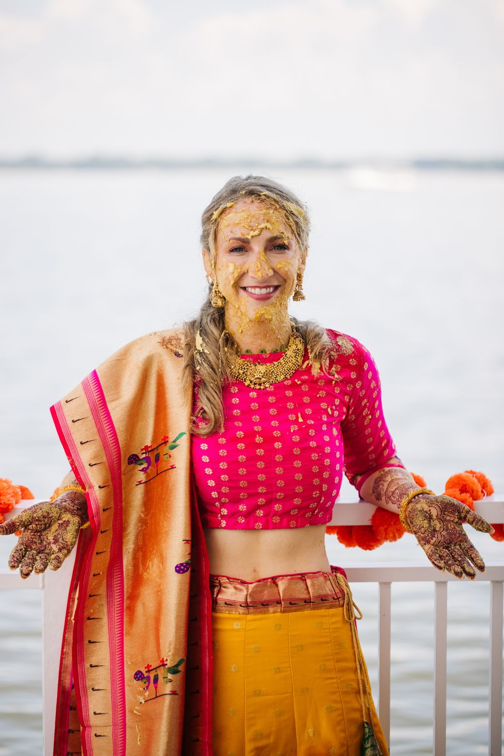 A joyful person in traditional Indian wedding attire with henna-decorated hands poses by the water wearing pink and orange.