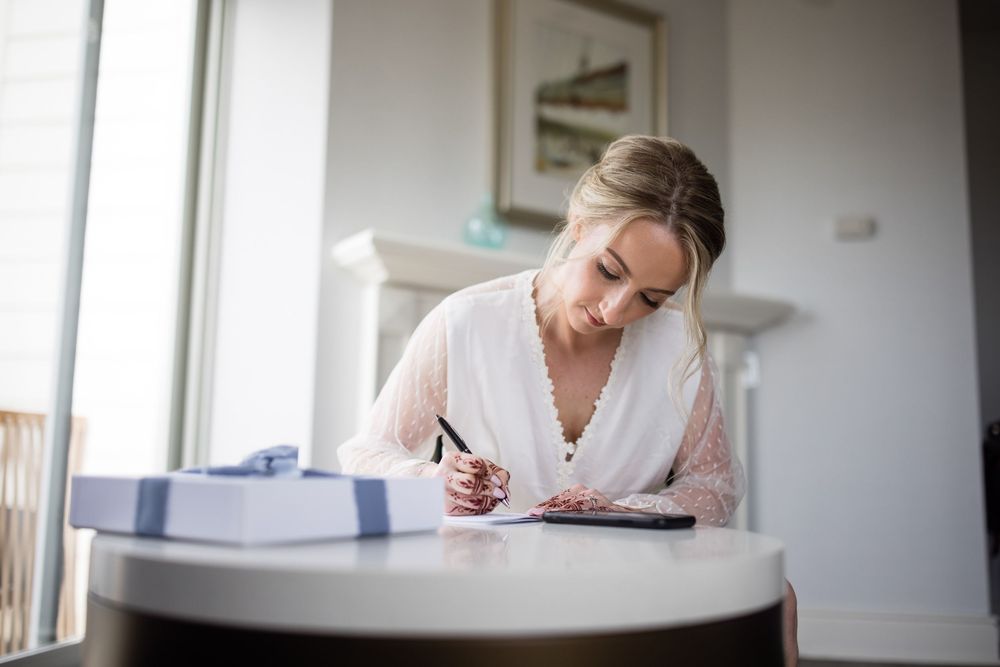 Someone in a white blouse writes notes while working at a round table near a bright window.