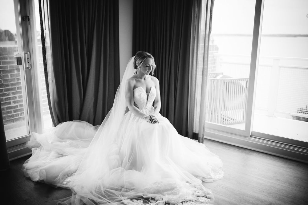 A black and white portrait of a bride in a flowing tulle gown sitting gracefully by a window.