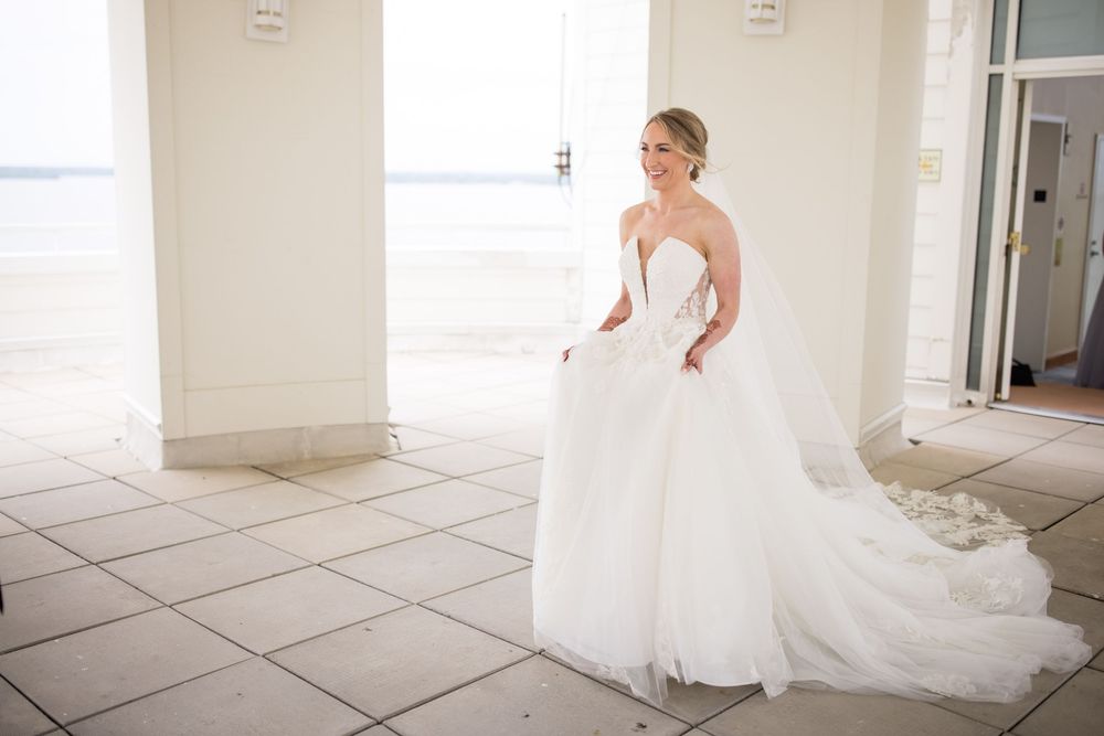 A bride in a strapless white wedding gown with a flowing train stands in a bright sunlit room with tiled floors.