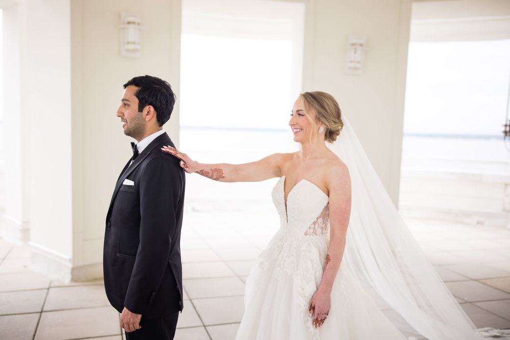 A bride in a white gown touches the shoulder of a groom in a black tuxedo during their first look wedding moment.