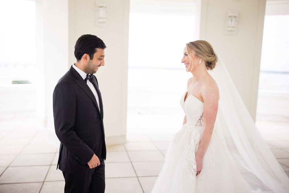 A bride in a white gown and veil shares a joyful first look moment with her groom in a black tuxedo.