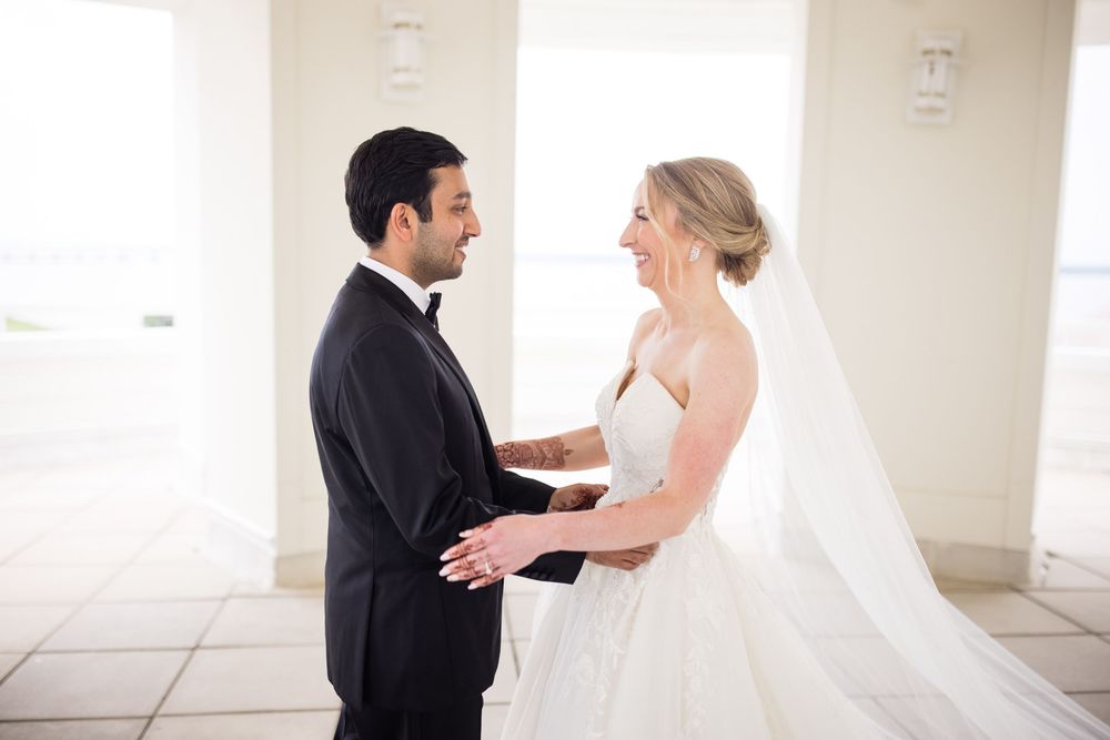 A bride in a white wedding gown and groom in black tuxedo share a tender first look moment indoors.