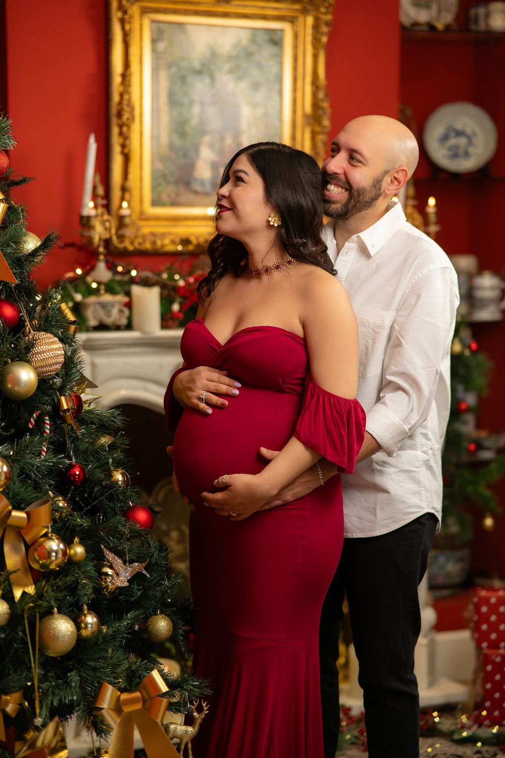 Romantic Christmas maternity photo session with couple embracing near decorated tree and red walls.