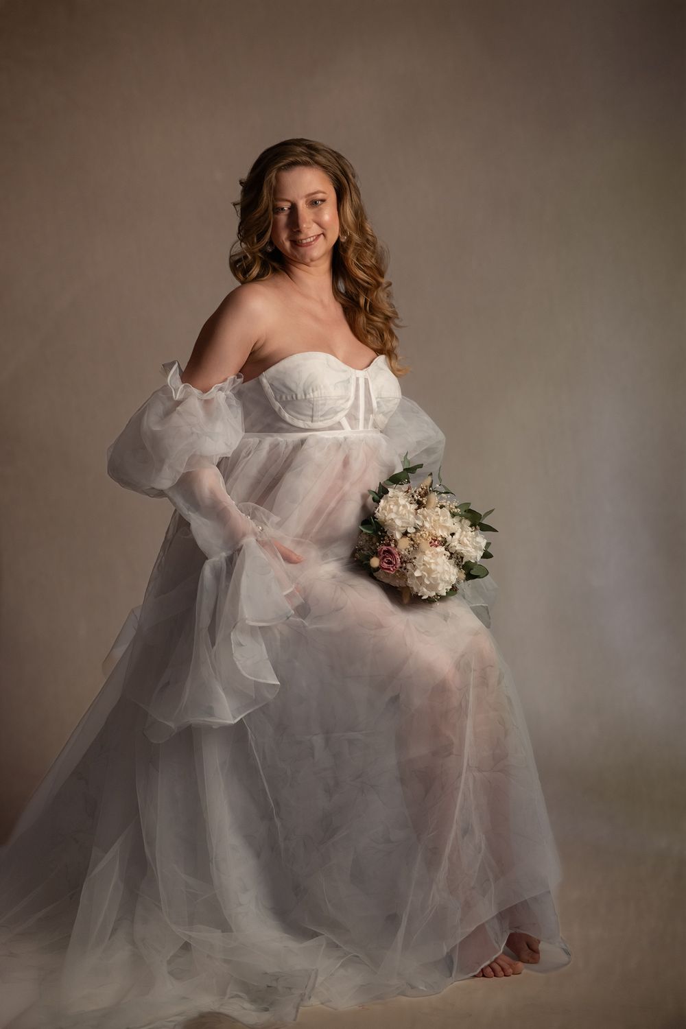 Ethereal bridal boudoir photo series showing a figure in flowing white tulle gown holding a delicate bouquet against neutral backdrop.