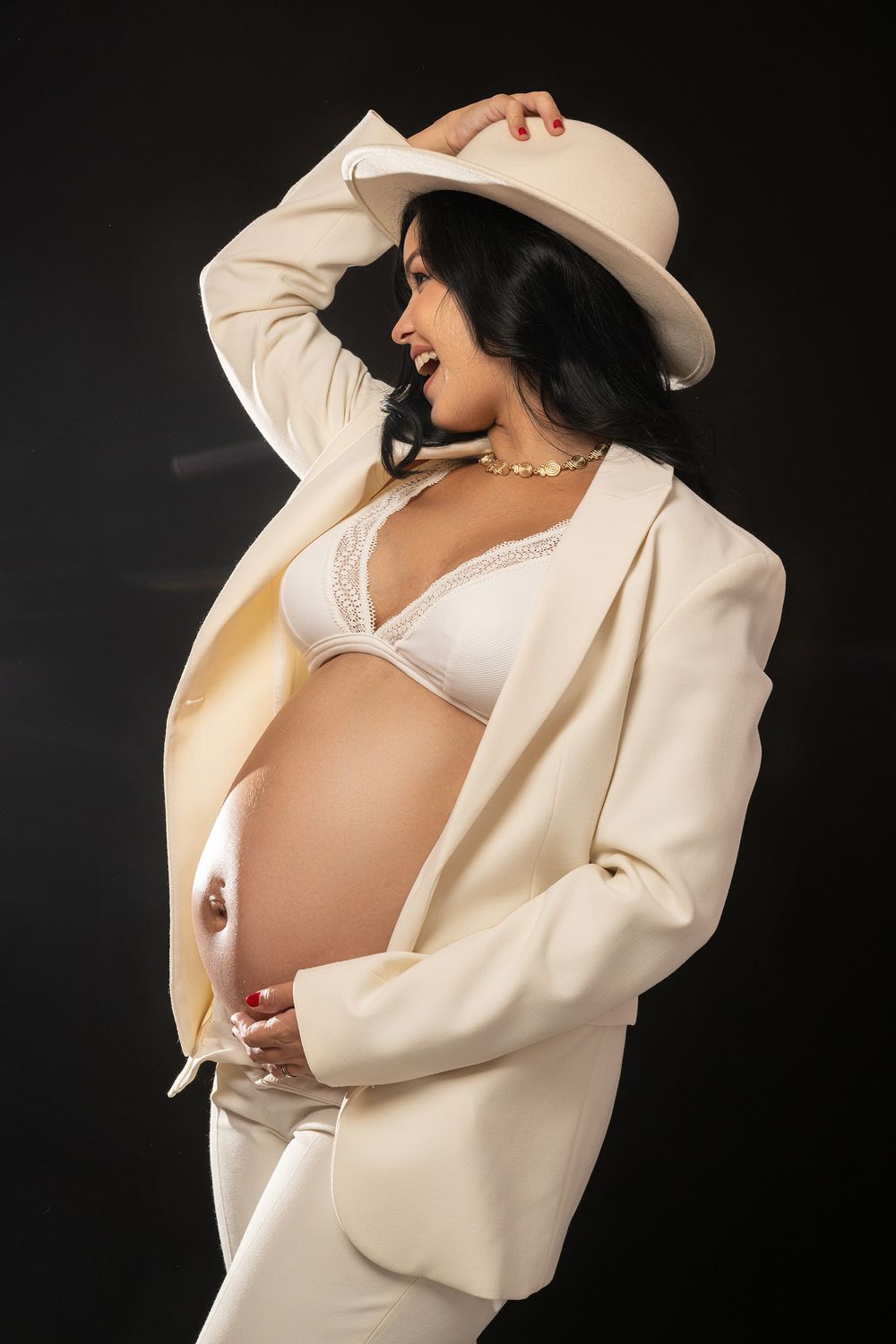 Elegant maternity portrait featuring cream colored outfit and hat with dramatic lighting against dark background.