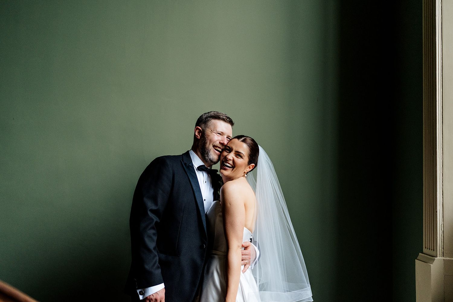 A bride and groom share a tender moment together while laughing against a green wall in an elegant wedding portrait.