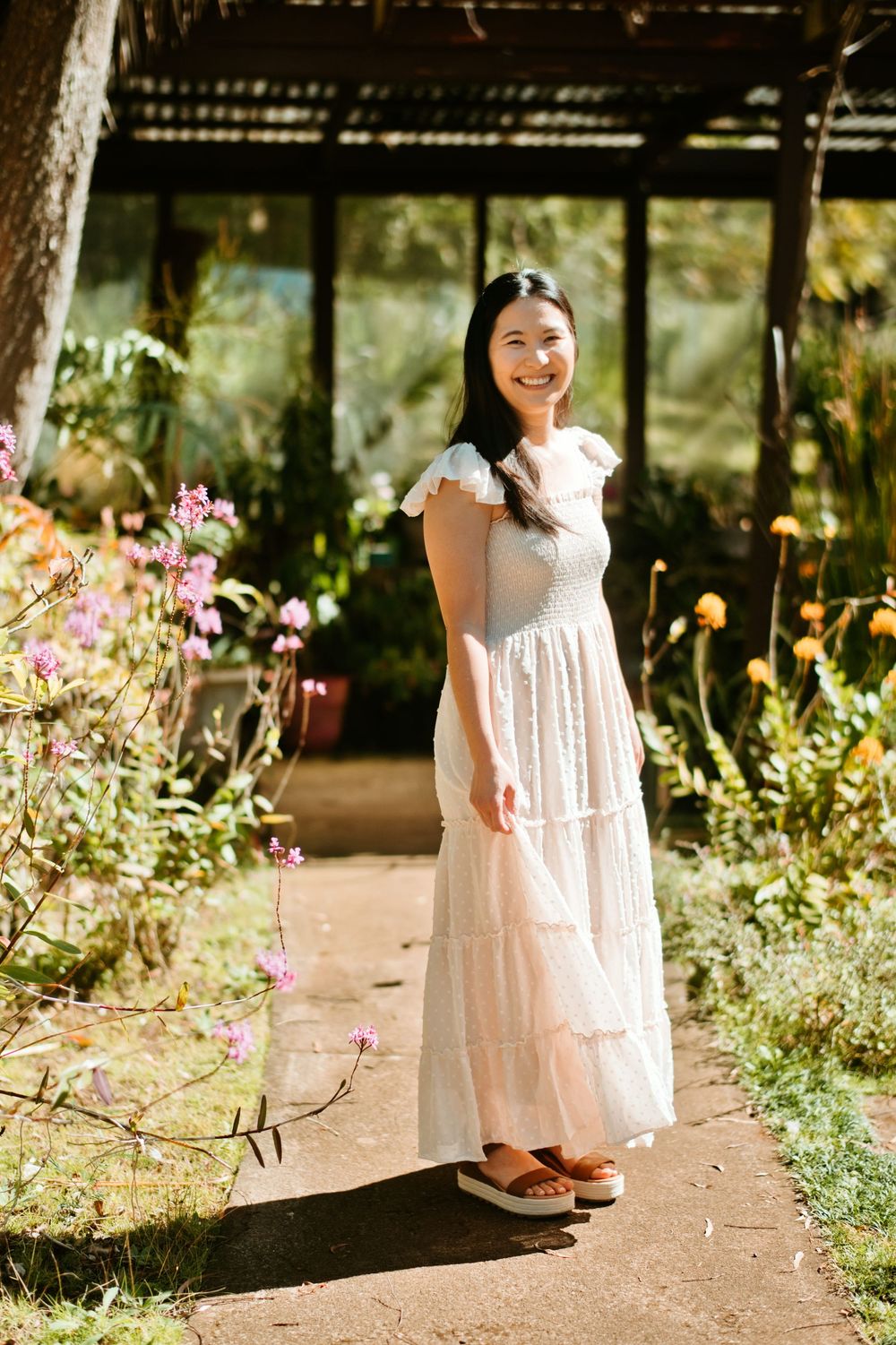 A person in a white dress stands on a garden path surrounded by blooming flowers and greenery on a sunny day.