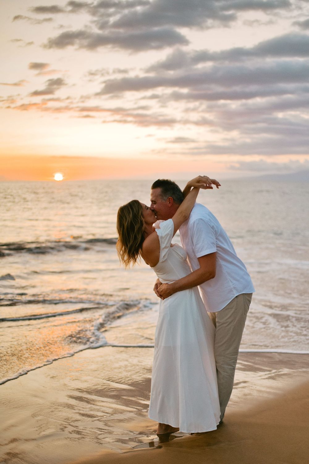 Couple embracing on sandy beach during golden hour sunset with ocean waves in the background.
