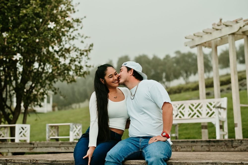 A couple shares a tender kiss by a white fence on a sunny afternoon.