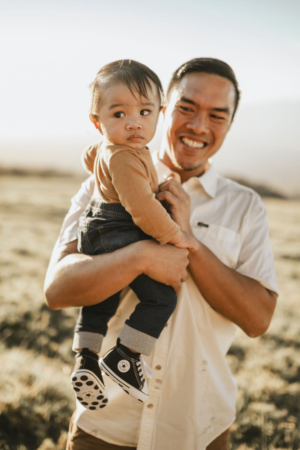 A heartwarming outdoor portrait of parent and toddler embracing in golden sunlight on a grassy hillside.