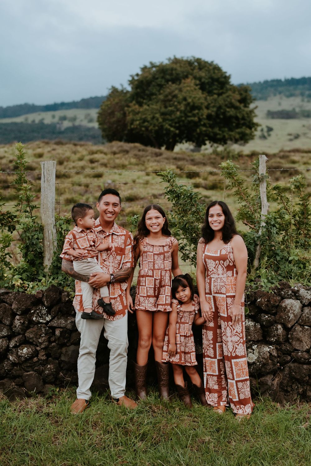 A group wearing matching terracotta-colored outfits poses together against a rustic stone wall in a pastoral setting.