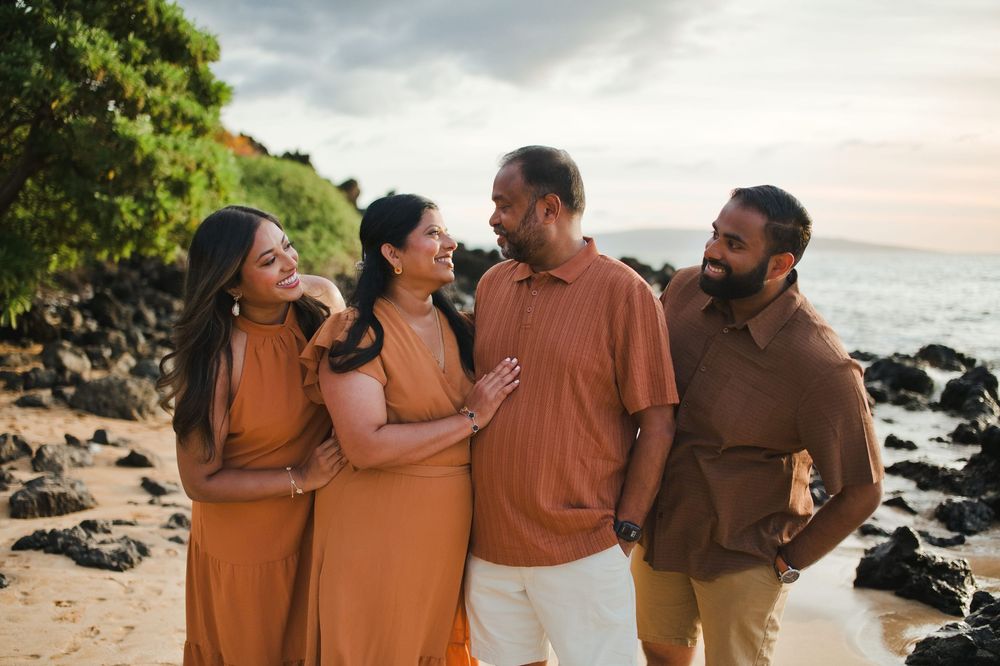 Group of friends laughing together on a beach at sunset with ocean and rocks in background.