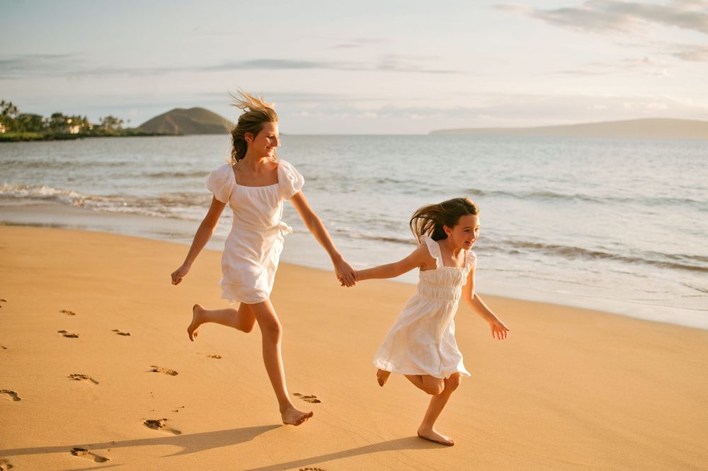 A couple in white outfits walks hand in hand along a golden sandy beach at sunset.