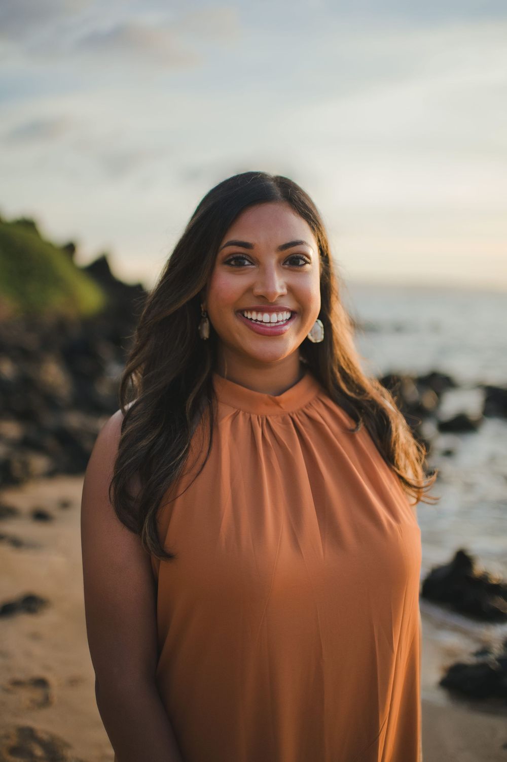 A woman in a sleeveless orange top smiles warmly while standing on a beach at sunset with rocks and ocean in background.