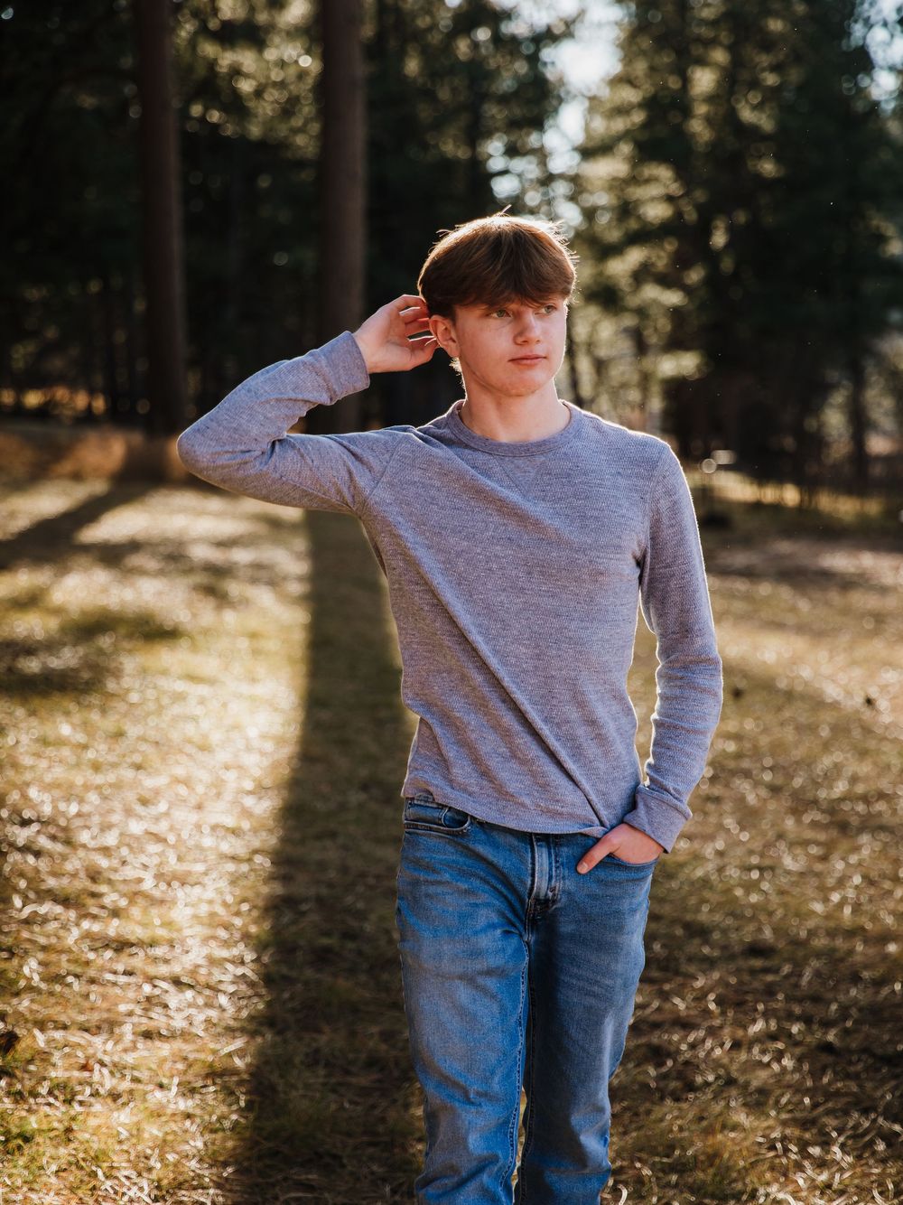 Person in grey sweater and jeans posing playfully in wooded outdoor setting.